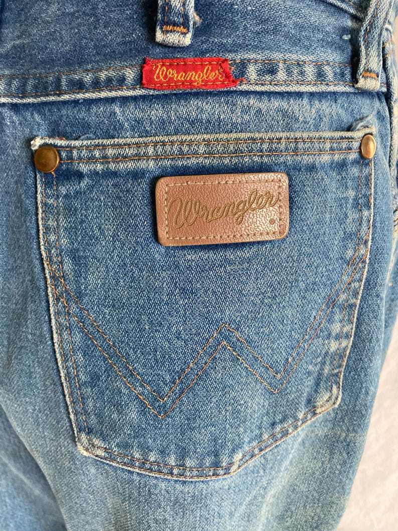 Pu&ograve; includere: Primo piano di jeans blu con una tasca posteriore con una toppa in pelle marrone e il nome del marchio "Wrangler" in rilievo. I jeans hanno un'etichetta rossa con il logo "Wrangler" e rivetti in ottone.