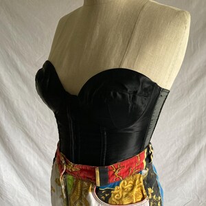 May include: A black satin corset with boning and a sweetheart neckline. The corset is worn over a pair of white denim jeans with a colorful patterned waistband.