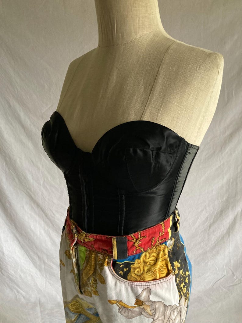 May include: A black satin corset with boning and a sweetheart neckline. The corset is worn over a pair of white denim jeans with a colorful patterned waistband.