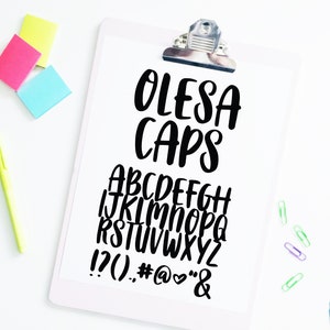 May include: A white clipboard with a black clip holds a sheet of paper with a handwritten font alphabet in black. The font is called "Olesa Caps" and includes uppercase letters, numbers, and punctuation marks. The background is white.