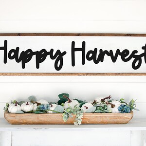 May include: A wooden sign with the text "Happy Harvest" in black script font. The sign is hanging on a white wall above a wooden bowl filled with white pumpkins and green foliage.
