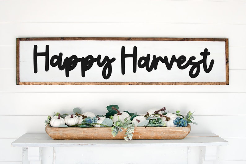 May include: A wooden sign with the text "Happy Harvest" in black script font. The sign is hanging on a white wall above a wooden bowl filled with white pumpkins and green foliage.