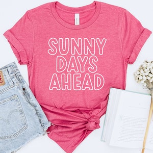以下が含まれることがあります： 白い輪郭のある文字で「SUNNY DAYS AHEAD」と書かれたピンクのTシャツです。