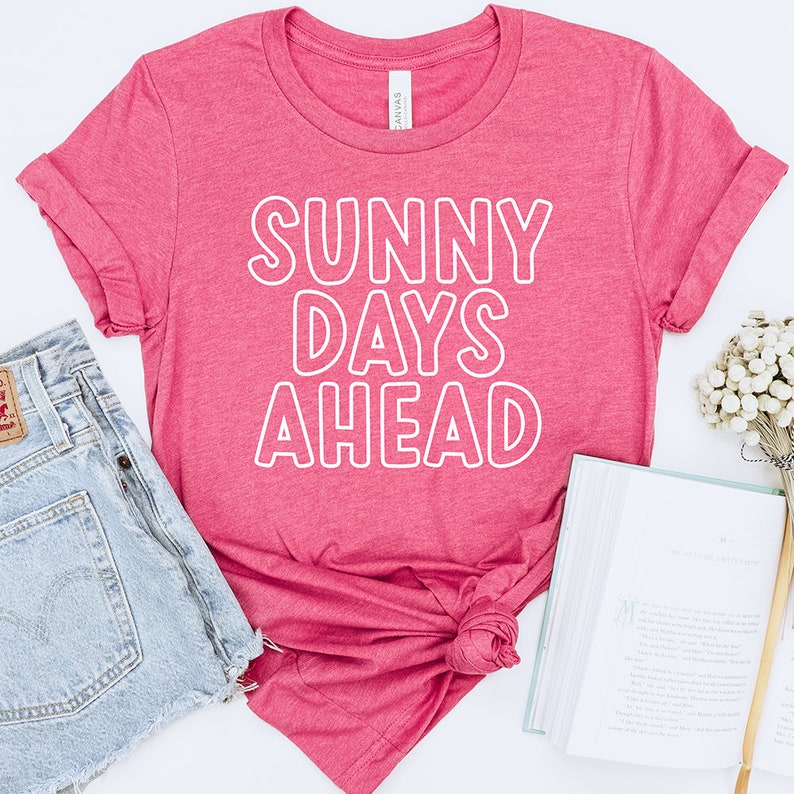 以下が含まれることがあります： 白い輪郭のある文字で「SUNNY DAYS AHEAD」と書かれたピンクのTシャツです。