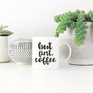May include: White ceramic coffee mug with black text that reads "but first, coffee". The mug is sitting on a white surface with two small potted succulents and a black and white patterned bowl.