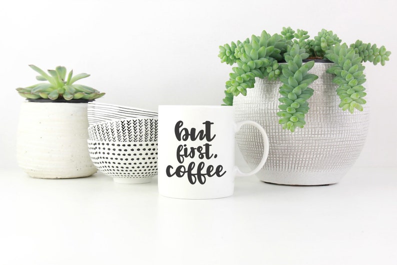 May include: White ceramic coffee mug with black text that reads "but first, coffee". The mug is sitting on a white surface with two small potted succulents and a black and white patterned bowl.