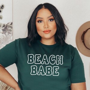 以下が含まれることがあります： 白い輪郭のある文字で「BEACH BABE」と書かれた濃い緑色のTシャツです。