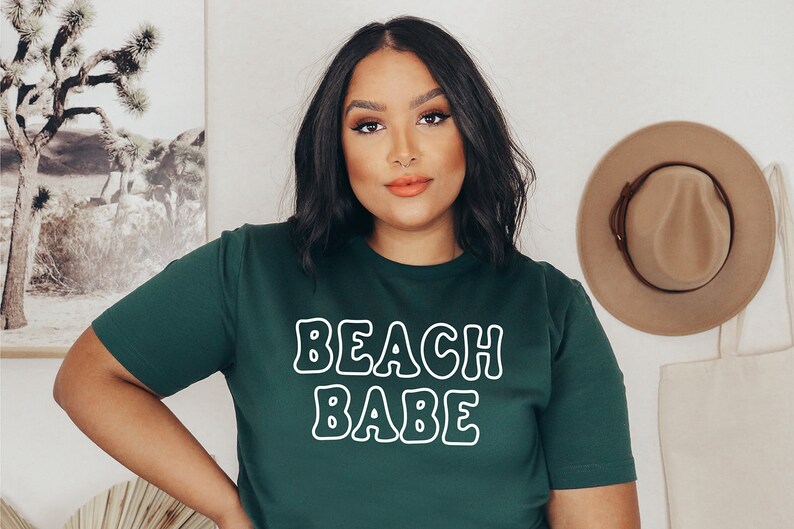 以下が含まれることがあります： 白い輪郭のある文字で「BEACH BABE」と書かれた濃い緑色のTシャツです。
