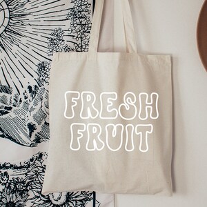 以下が含まれることがあります： 白いキャンバス地のトートバッグに、白い輪郭の文字で「FRESH FRUIT」と印刷されています。