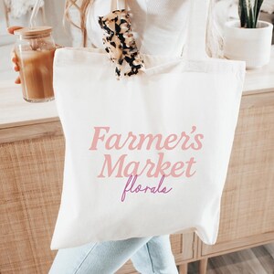 May include: A white canvas tote bag with the text "Farmer's Market florals" in pink and purple. The bag is held in front of a wooden cabinet. A glass jar with a straw and a drink is visible.