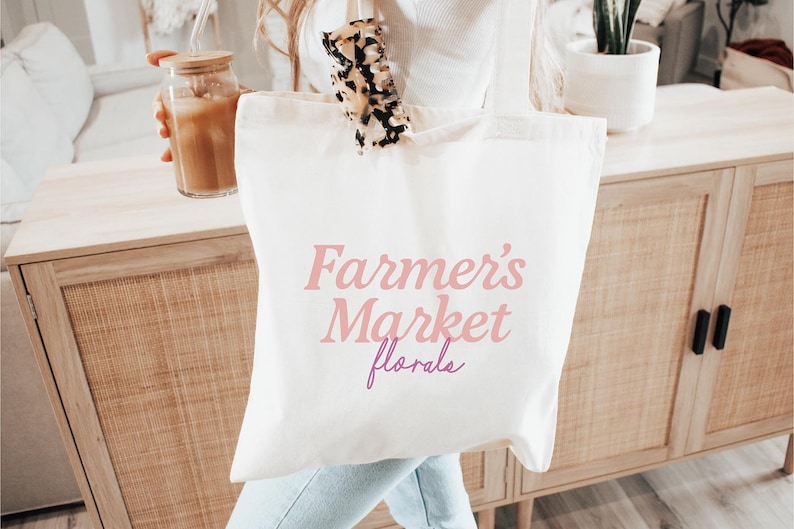 May include: A white canvas tote bag with the text "Farmer's Market florals" in pink and purple. The bag is held in front of a wooden cabinet. A glass jar with a straw and a drink is visible.