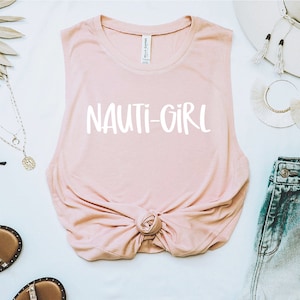 May include: A pink sleeveless muscle tank top with the text "NAUTI-GIRL" printed in white on the front. The top is knotted at the bottom and is styled with denim shorts, a straw hat, and jewellery.