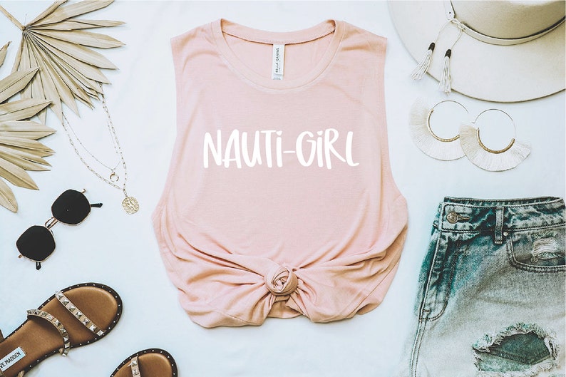 May include: A pink sleeveless muscle tank top with the text "NAUTI-GIRL" printed in white on the front. The top is knotted at the bottom and is styled with denim shorts, a straw hat, and jewellery.