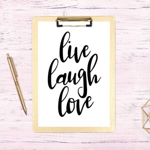 May include: A white clipboard with a gold clip holds a white sheet of paper with the words "live laugh love" written in black cursive script. A gold pen and a gold candle holder are on a pink wooden surface.