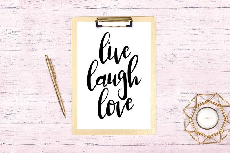 May include: A white clipboard with a gold clip holds a white sheet of paper with the words "live laugh love" written in black cursive script. A gold pen and a gold candle holder are on a pink wooden surface.