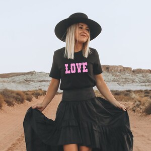 Puede incluir: Una mujer con un sombrero negro de ala ancha, una camiseta negra con la palabra "LOVE" en letras rosas y una falda negra de capas. Est&aacute; de pie en un paisaje des&eacute;rtico.