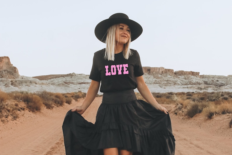 Puede incluir: Una mujer con un sombrero negro de ala ancha, una camiseta negra con la palabra "LOVE" en letras rosas y una falda negra de capas. Est&aacute; de pie en un paisaje des&eacute;rtico.