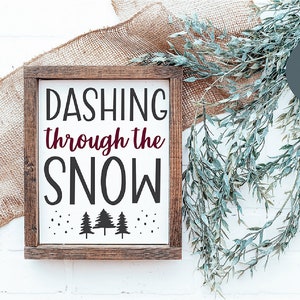 Pu&ograve; includere: Un cartello di legno bianco con il testo "Dashing through the snow" in lettere nere. Il cartello &egrave; decorato con piccoli fiocchi di neve neri e tre alberi di pino neri in silhouette. Il cartello &egrave; incorniciato in legno marrone.