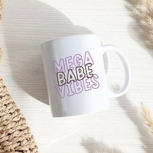以下が含まれることがあります： 白いセラミック製のマグカップに、紫色の輪郭で「MEGA BABE VIBES」という文字が重ねて書かれています。