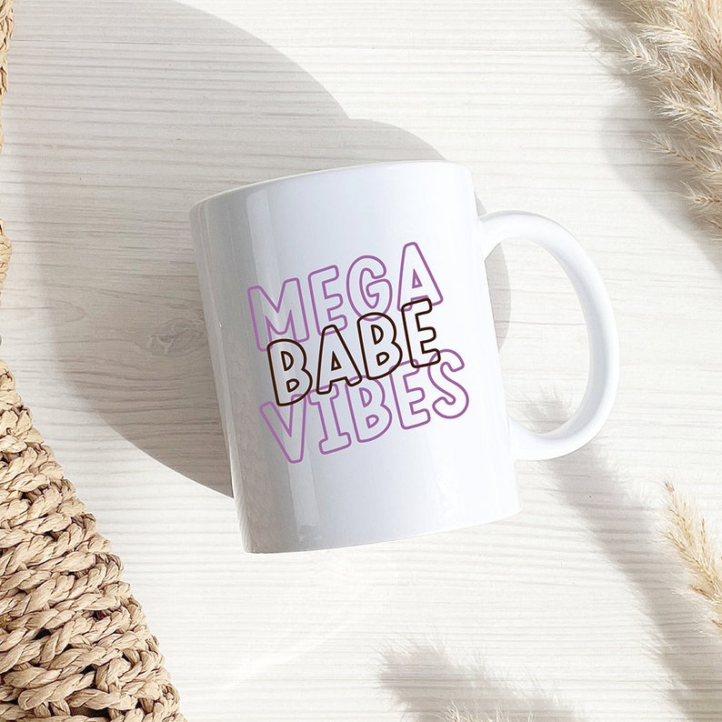 以下が含まれることがあります： 白いセラミック製のマグカップに、紫色の輪郭で「MEGA BABE VIBES」という文字が重ねて書かれています。