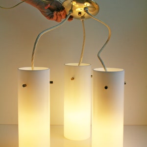 May include: Three white cylindrical pendant lights with brass accents hang from a ceiling fixture. The lights are illuminated and cast a warm glow.