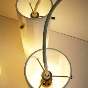May include: Two white cylindrical lampshades with a white cord connecting them. The cord is curved and the lampshades are lit.