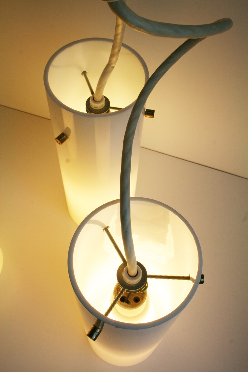 May include: Two white cylindrical lampshades with a white cord connecting them. The cord is curved and the lampshades are lit.