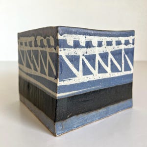 May include: A square ceramic container with a blue and white geometric design. The top section features a white pattern on a blue background, with a black band in the middle. The bottom edge is a lighter blue.