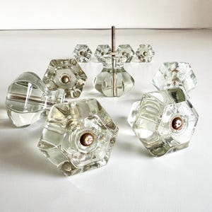 May include: Clear glass cabinet knobs with a hexagonal design and brass-colored hardware. The knobs are arranged on a white surface, showcasing their faceted surfaces and vintage aesthetic. A set of vintage cabinet hardware.