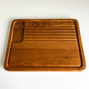 May include: A rectangular wooden cutting board with a warm brown finish and visible wood grain. The board features a smooth flat surface, a deep recessed rectangular section on the left, and five horizontal grooves on the upper right side.