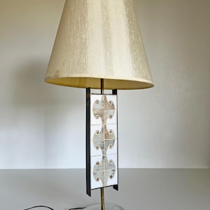 May include: A vintage table lamp with a cream-colored lampshade. The lamp features a black metal frame with decorative tiles in shades of gray and white. The base is a brushed silver oval, and the lamp has a cord.
