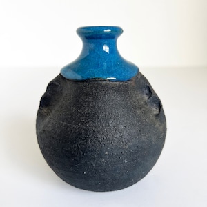 May include: A small, round, black ceramic vase with a blue glazed top. The vase has a rough, textured surface.