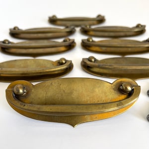 May include: A collection of antique brass drawer pulls. Each pull has a curved, oval shape with a decorative ball on each end. The brass has a warm, aged patina, suggesting vintage or reclaimed hardware. The pulls are arranged on a white surface.