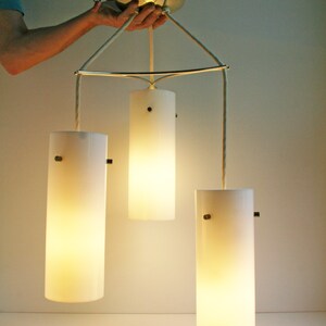May include: A three-light pendant light fixture with white frosted glass shades. The shades are cylindrical and hang from a metal frame.