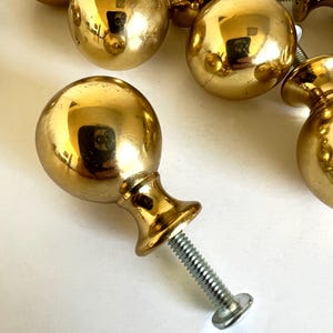 May include: A collection of shiny, gold-colored cabinet knobs. Each knob has a round, ball-shaped top and a decorative base, attached to a threaded screw. The knobs are reflective, with a polished finish, and are arranged on a white surface.
