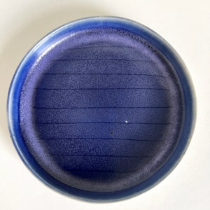 May include: A blue ceramic plate with a textured surface and a subtle gradient of color. The plate has a slightly raised rim and a smooth, glazed finish.