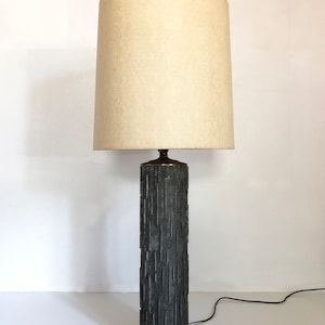 May include: A tall, gray, textured lamp with a white fabric shade. The lamp base is cylindrical and has a carved design. The lamp sits on a white marble base.