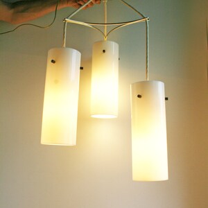 May include: A three-light pendant light fixture with white cylindrical shades. The fixture is suspended from the ceiling by three cords and has a gold metal frame.