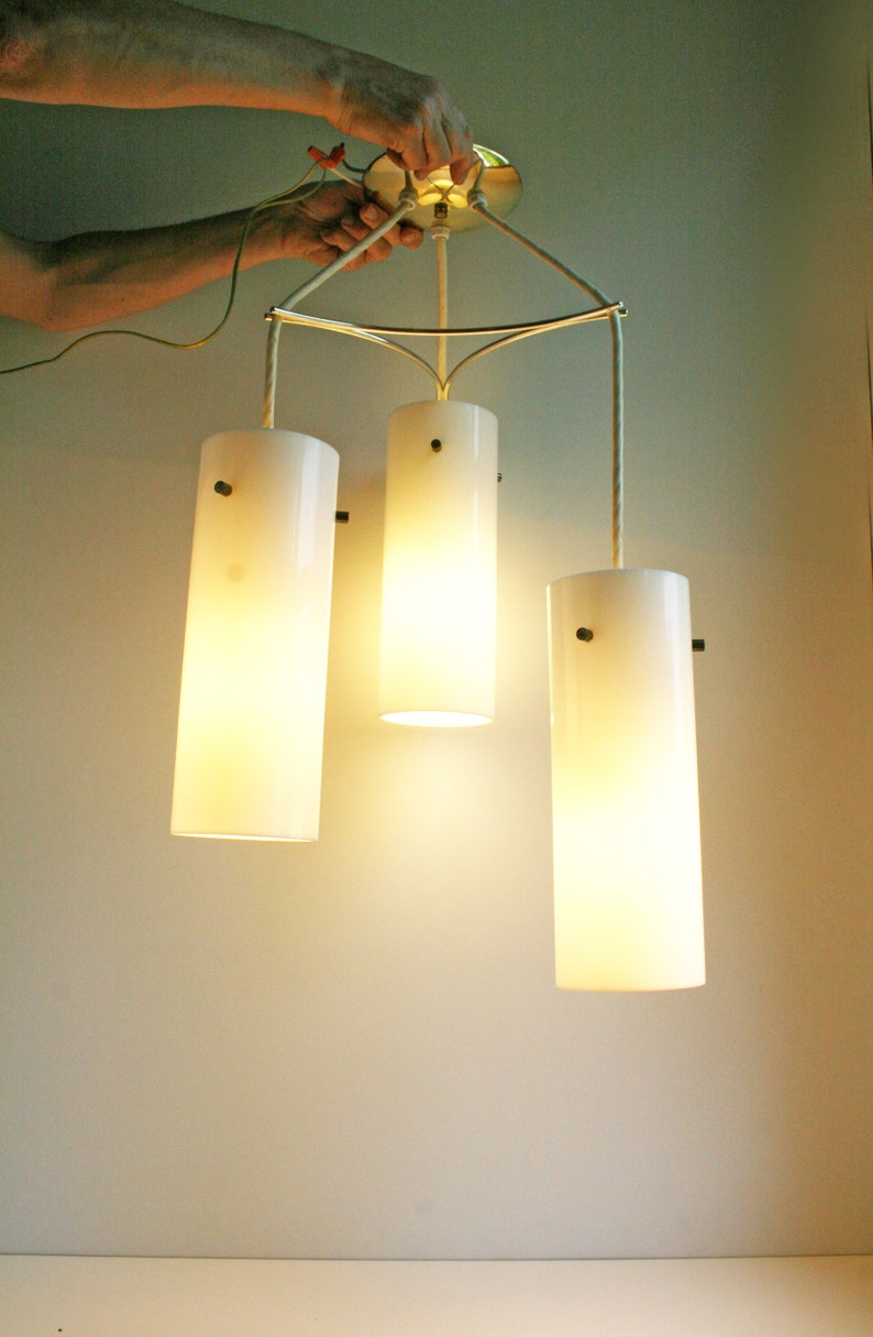 May include: A three-light pendant light fixture with white cylindrical shades. The fixture is suspended from the ceiling by three cords and has a gold metal frame.