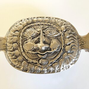 May include: Silver-colored, oval-shaped decorative object with a detailed face design. The face has prominent features, including eyes, a nose, and a smiling mouth. The object has two handles on either side and is likely a decorative piece.