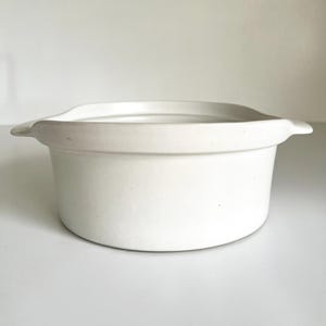 May include: A white, round ceramic baking dish with a slightly flared rim and two small handles. The dish has a smooth, matte finish and is suitable for oven use. The overall design is simple and functional, perfect for baking and serving.