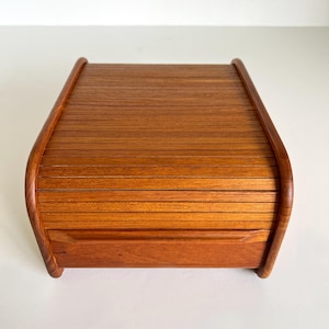 May include: A wooden roll-top box with a warm, reddish-brown finish. The box features a curved top and sides, with horizontal wooden slats forming the roll-top. The box is rectangular with a small drawer at the front.