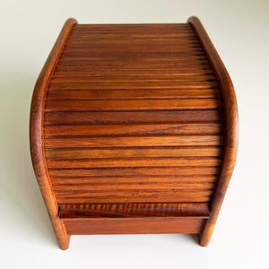 May include: A medium-brown wooden bread box featuring a curved top with a slatted roll-top design. The structure is composed of horizontal wooden slats that form a rounded lid, supported by two curved side panels that extend down to form small feet.