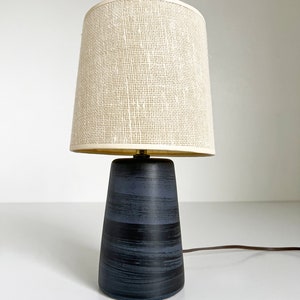 May include: A black ceramic table lamp with a white woven lampshade. The lamp has a conical base with a brushed black finish.