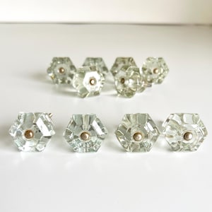 May include: A collection of clear, hexagonal glass cabinet knobs with a silver-colored base. The knobs are arranged in two rows, with a total of eight knobs visible. The glass has a slight green tint.