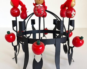May include: A black metal candle holder with a circular base and six arms. Each arm has a red wooden apple and a red wooden doll with a painted face and a red hat.