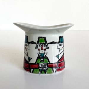 May include: White ceramic vase shaped like a hat with a geometric design of three stylized faces in black, red, green, and blue.