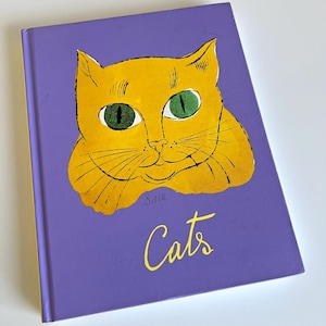 May include: A hardcover book titled "Cats" with a vibrant illustration of a yellow cat on a purple background. The cat has green eyes and the title is in yellow script. The book is likely a children's book or a book about cats.