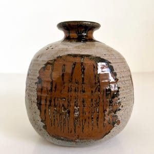 May include: A small, round ceramic vase with a brown and white glaze. The vase has a wide mouth and a narrow neck. The glaze is uneven, creating a mottled effect.