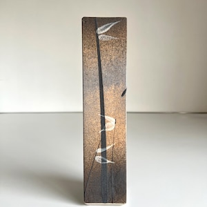 May include: A rectangular ceramic vase with a textured surface. The vase features a design of tall, thin black lines and white leaf-like shapes against a gradient of brown and gray tones. The vase is standing upright.